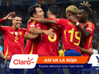 Spain national team players celebrate a goal, embracing in red jerseys on the field with a cheering crowd in the background.