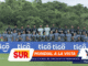 Group of soccer players posing for a team photo on the field, gray/black kits, with blue 'tigo' banners in front.