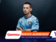 Portrait of a man in a light-blue jersey with arms crossed, standing against a dark gradient backdrop with colorful graphic overlays at the left and bottom edges, including sponsor logos.