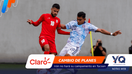 Two soccer players clash for the ball during a match: one in a red kit (#10), the other in a light blue patterned kit (#20) near a yellow post.