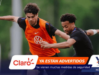 Two male soccer players in action during a match, one in an orange training vest chasing the ball, with sponsor banners across the bottom of the image.