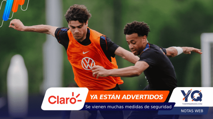 Two male soccer players in action during a match, one in an orange training vest chasing the ball, with sponsor banners across the bottom of the image.