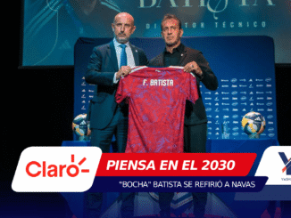 Two men in suits hold up a red patterned soccer jersey with the name F. Batista on stage at a press event, with blue sponsor banners in the background.