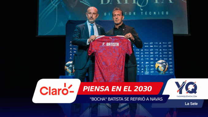 Two men in suits hold up a red patterned soccer jersey with the name F. Batista on stage at a press event, with blue sponsor banners in the background.
