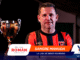 Portrait of a man in a red-and-black striped soccer jersey standing beside a silver trophy; sponsor banners cover the lower part of the image.