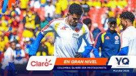 Colombia soccer player in a white jersey with crest and blue sleeves, amid a colorful stadium crowd; banner shows Claro and 'EN GRAN NIVEL' for Mundial 2026