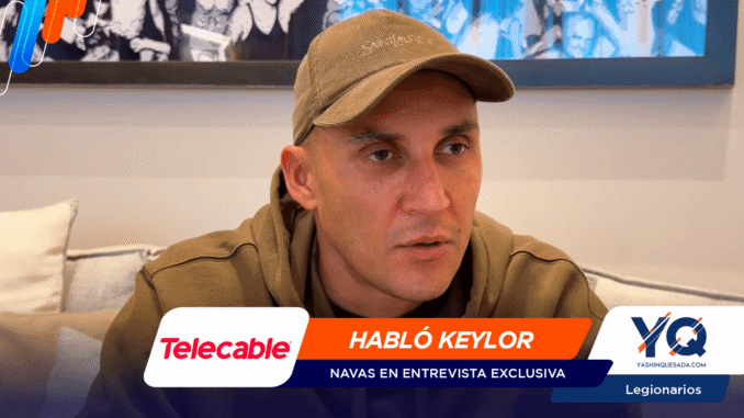 Man in a brown hoodie and tan cap sits on a sofa during an interview thumbnail, with an orange banner 'HABLÓ KEYLOR' and sponsor logos (Telecable, YQ).