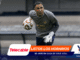 Soccer goalkeeper in gray shirt and white gloves reaches for a ball during training, blurred stadium seats in the background.