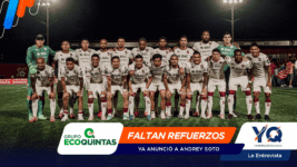 Soccer team in white uniforms posing for a group photo on a night-lit field, players standing and kneeling together with two green-clad goalkeepers at each end.
