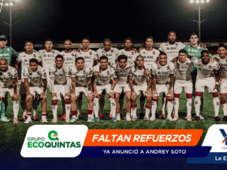 Soccer team in white uniforms posing for a group photo on a night-lit field, players standing and kneeling together with two green-clad goalkeepers at each end.