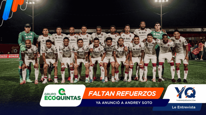 Soccer team in white uniforms posing for a group photo on a night-lit field, players standing and kneeling together with two green-clad goalkeepers at each end.