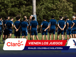 Soccer players in blue uniforms standing in a circle on a grassy field as a coach talks to them; foreground shows sponsor banners overlapping the bottom edge.