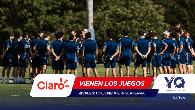 Soccer players in blue uniforms standing in a circle on a grassy field as a coach talks to them; foreground shows sponsor banners overlapping the bottom edge.
