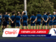 Soccer players in blue uniforms standing in a circle on a grassy field as a coach talks to them; foreground shows sponsor banners overlapping the bottom edge.