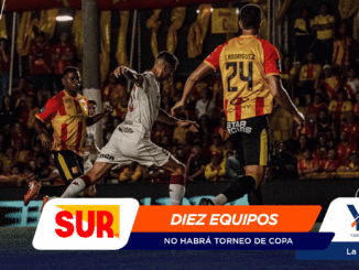 Soccer players in yellow-red and white jerseys compete for the ball on a crowded stadium field, with colorful sponsor banners at the bottom.