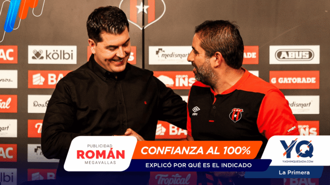 Two men shake hands on a stage with sponsor banners behind them, smiling at each other.