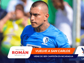 Athlete in a blue soccer jersey during a match, tattoos on the neck, with sponsor banners in the foreground reading 'VUELVE A SAN CARLOS' and 'ROMÁN'.
