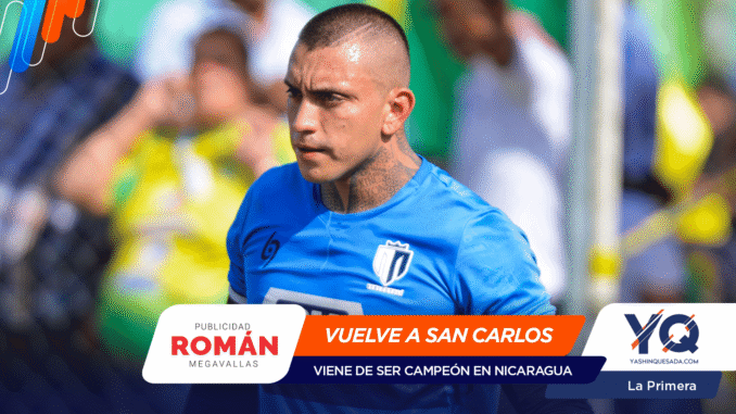 Athlete in a blue soccer jersey during a match, tattoos on the neck, with sponsor banners in the foreground reading 'VUELVE A SAN CARLOS' and 'ROMÁN'.