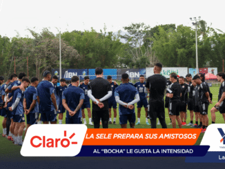 Soccer players in blue and black gear form a huddle on a green training field as coaches speak, with banners and floodlights in the background.