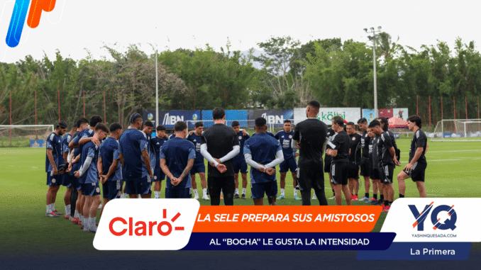 Soccer players in blue and black gear form a huddle on a green training field as coaches speak, with banners and floodlights in the background.