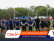 Soccer players in blue and black gear form a huddle on a green training field as coaches speak, with banners and floodlights in the background.