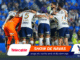Soccer team huddling in a blue stadium, players in white and navy uniforms with orange goalie, banners at bottom display sponsors like Telecable and YQ Vivans.