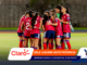Women's soccer team huddled together in red uniforms on a grassy field before a match, showing teamwork and unity.