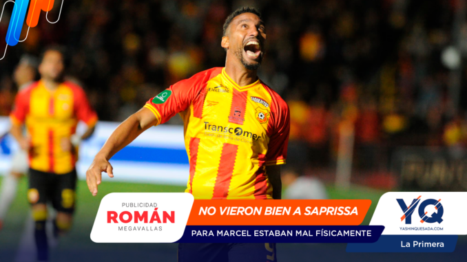 Soccer player in red and yellow jersey celebrates with mouth open on a blurred stadium background; foreground has sponsor banners.