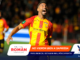 Soccer player in red and yellow jersey celebrates with mouth open on a blurred stadium background; foreground has sponsor banners.