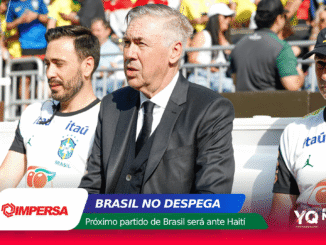 Man in a dark suit sits among Brazil football players at a team event, with a colorful banner and crowd behind him