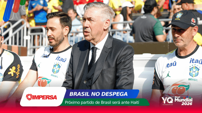 Man in a dark suit sits among Brazil football players at a team event, with a colorful banner and crowd behind him