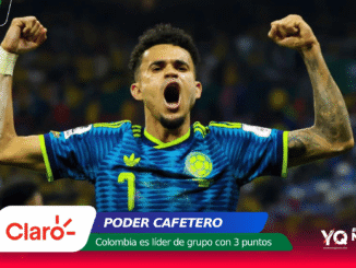 Colombian soccer player in blue jersey raises fists in celebration on a stadium backdrop with sponsor banners at the bottom.