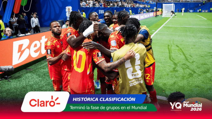 Group of soccer players in red uniforms celebrate in a tight huddle on the pitch near the sideline; one wears a gold vest with 'FIFA World Cup 2026' on the back.