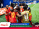Group of soccer players in red uniforms celebrate in a tight huddle on the pitch near the sideline; one wears a gold vest with 'FIFA World Cup 2026' on the back.