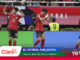 Three male players in red jerseys celebrate on an indoor futsal court as photographers watch in the background, with sponsor banners nearby.