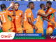 Ivory Coast football players in orange jerseys celebrate together on the field with a blue stadium backdrop and sponsor graphics at the bottom.
