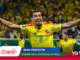 Colombian soccer player in a yellow jersey with arms outstretched, celebrating on the field with a cheering crowd in Miami behind him.