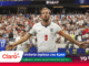 England striker Harry Kane celebrates a goal with arms outstretched as a full stadium cheers behind him, wearing England white kit with number 9