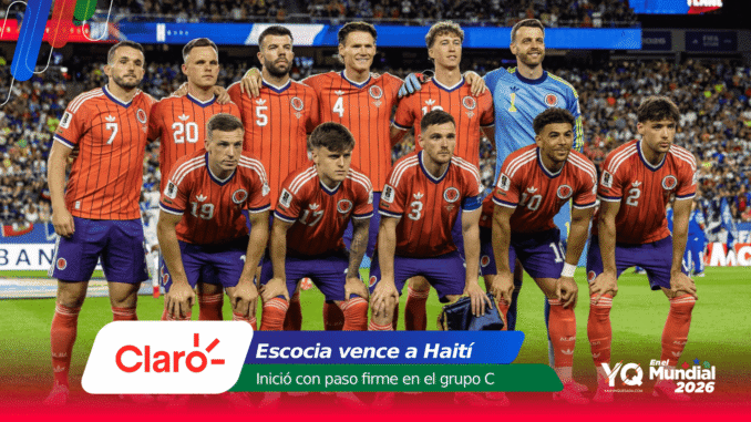 Group photo of the Colombian national football team posing on the field before a match, in red jerseys and blue shorts, with a crowded stadium in the background.