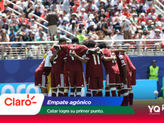 Soccer players in maroon uniforms huddle with arms around each other on the field as a large crowd watches from the stands behind them in the stadium scene.
