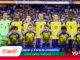 Swedish national football team players pose for a group photo on the pitch before a match, wearing yellow jerseys with blue trim; goalkeeper in green stands at left, stadium crowd in background.