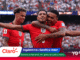 Three England footballers in red jerseys celebrate a goal on the pitch as a packed stadium cheers in the background.