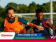 Two soccer players competing for the ball on a sunny field, one in an orange bib and the other in a dark jersey; banners overlay the bottom area.