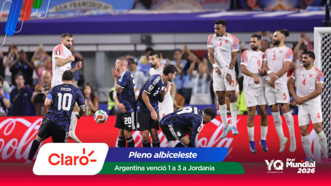 Soccer players in white uniforms stand near the goal as rivals in dark blue jerseys kick the ball midair during a match, with banners and sponsor logos in the foreground and a crowd in the stands behind.