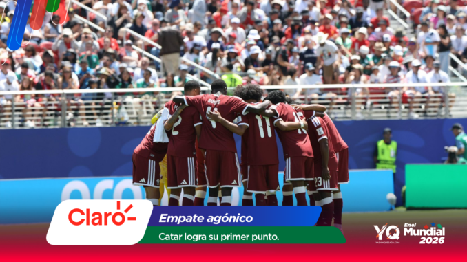 Soccer players in maroon uniforms huddle with arms around each other on the field as a large crowd watches from the stands behind them in the stadium scene.