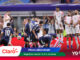 Soccer players in white uniforms stand near the goal as rivals in dark blue jerseys kick the ball midair during a match, with banners and sponsor logos in the foreground and a crowd in the stands behind.