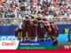 Soccer players in maroon uniforms huddle with arms around each other on the field as a large crowd watches from the stands behind them in the stadium scene.