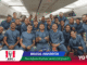 Group of athletes in matching blue outfits posing together inside an airplane cabin, smiling at the camera.