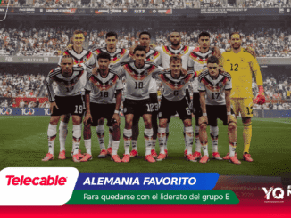 Germany national football team players in a group pose on the pitch, white kits with black-red-gold accents, goalkeeper in yellow, stadium crowd behind them