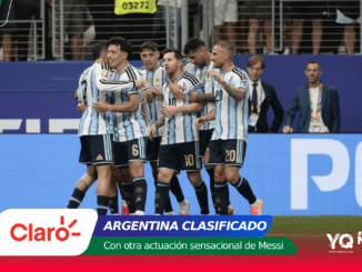 Argentina national team players celebrate together on the pitch after a victory, blue and white striped uniforms visible against a blue stadium barrier and banners in the foreground.